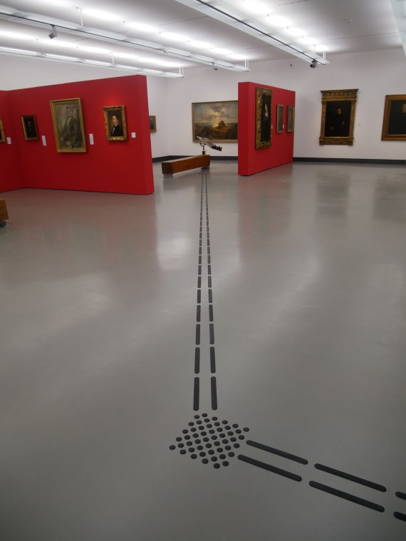 Ein Blindenleitsystem auf grauem Boden führt in einem Museumsraum zu einer Bank. An den Wänden hängen Gemälde.