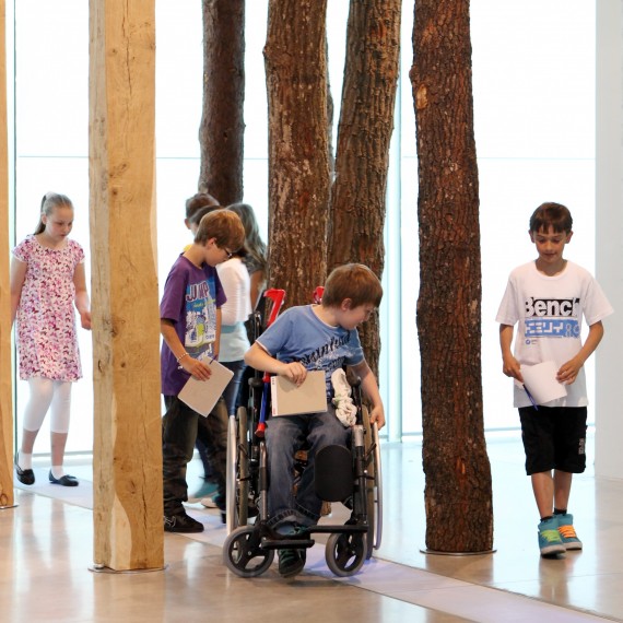 Eine Kindergruppe in einem Museum läuft zwischen aufgestellten Holzpfählen. Ein Kind sitzt im Rollstuhl.