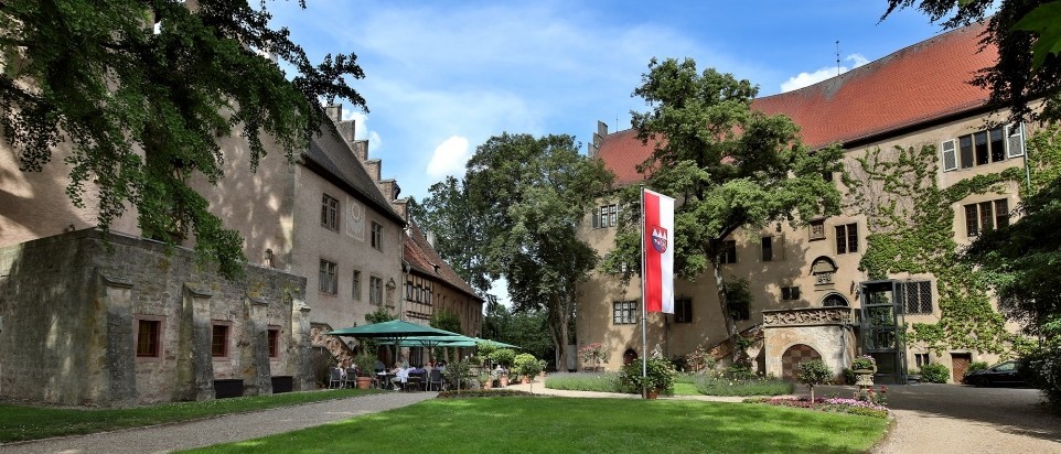 Um einen begrünten Hof gruppieren sich zwei historische Schlossgebäude. Das Hauptschloss ist dreistöckig. Vor dem kleineren Schloss stehen sind Sonnenschirme, Stühle und Tische.