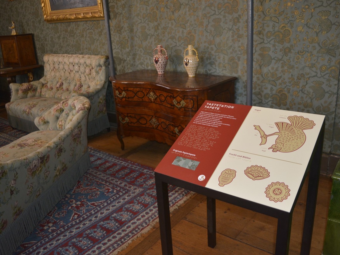 eine Tafel ist angeschrägt auf auf einem vierbeinigen Gestell aufgeschraubt. Im Hintergrund stehen eine Kommode und zwei historische Sofas.