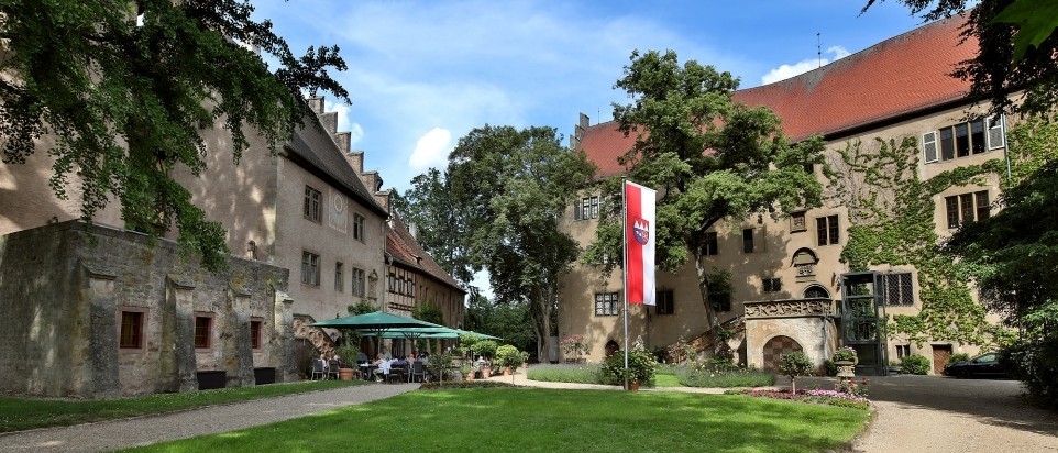Um einen begrünten Hof gruppieren sich zwei historische Schlossgebäude. Das Hauptschloss ist dreistöckig. Vor dem kleineren Schloss stehen sind Sonnenschirme, Stühle und Tische.