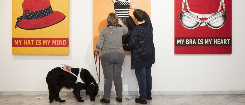 An einer Wand hängen drei Gemälde. Zwei Personen berühren das tastbares Gemälde in der Mitte. Neben ihnen steht ein Blindenführhund.