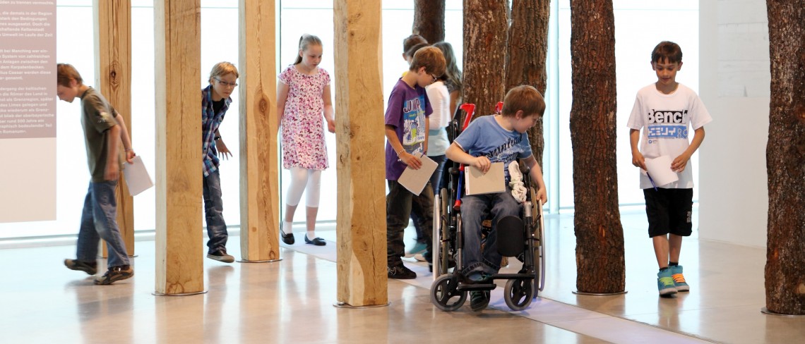 Eine Kindergruppe in einem Museum läuft zwischen aufgestellten Holzpfählen. Ein Kind sitzt im Rollstuhl.