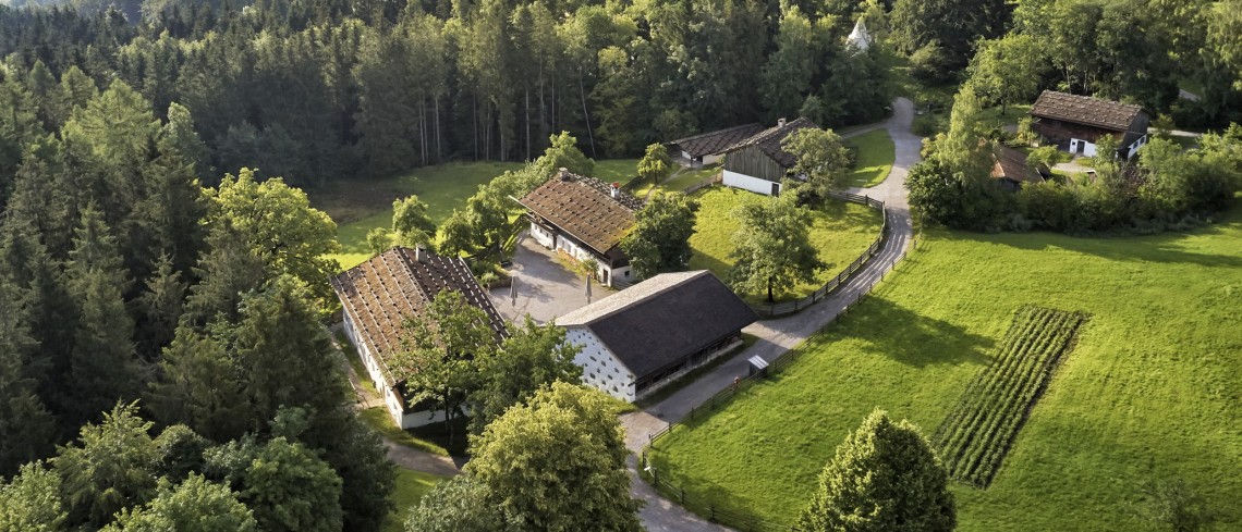 Eingebettet in grüne Wiese und Wald sind aus der Vogelperspektive ein historischer Bauernhof mit verschiedenen Nebengebäuden.