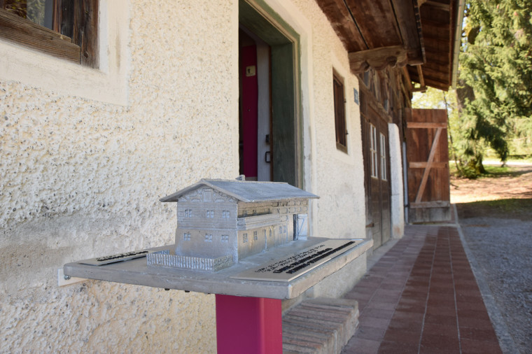 Ein tastbares Modell eines Bauernhauses steht auf einer Stele vor dem Eingang eines Hauses.