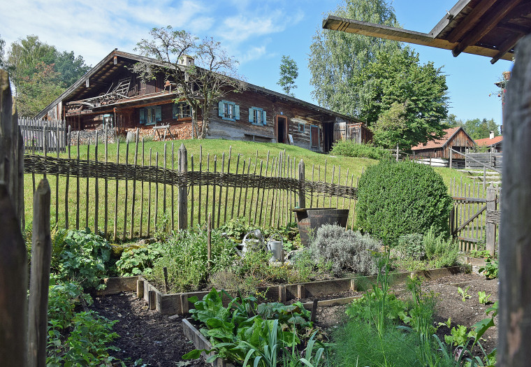 Ein historischer Bauerngarten mit Beeten voller Gem&uuml;se. Der Garten ist mit einem Zaun gesichert. Im Hintergrund steht ein Holzhaus auf einem H&uuml;ge.