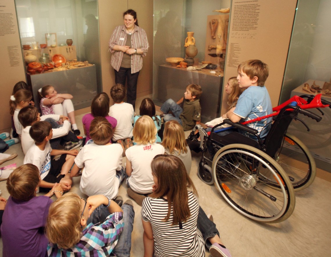 Vor zwei Vitrinen mit römischen Objekten steht eine Museumsführerin. Vor ihr sitzt eine Gruppe von Kindern auf dem Boden. Ein Kind sitzt im Rollstuhl.