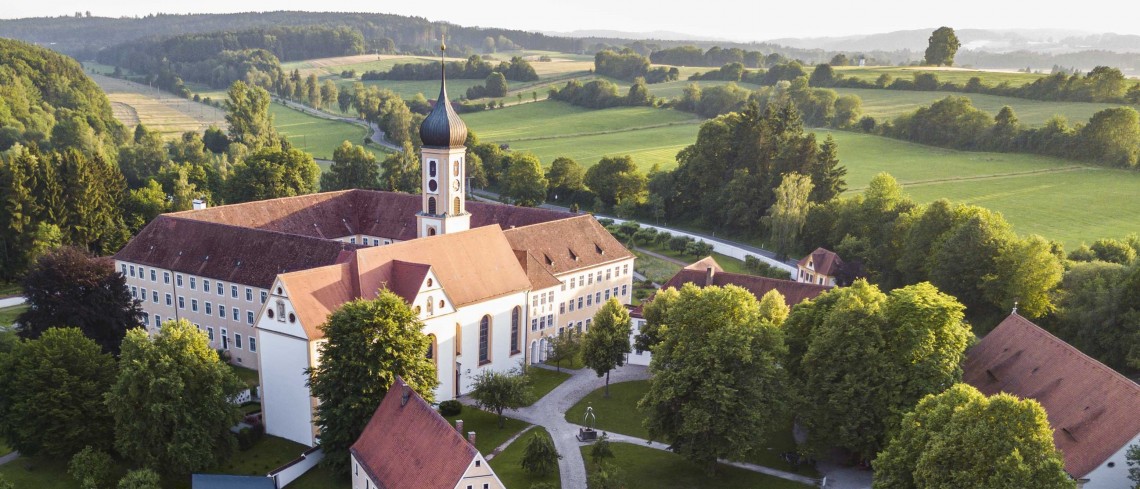 Ein Klostergebäude mit Kirche und Zwiebelturm steht in einer Landschaft mit Wiesen und Hecken.