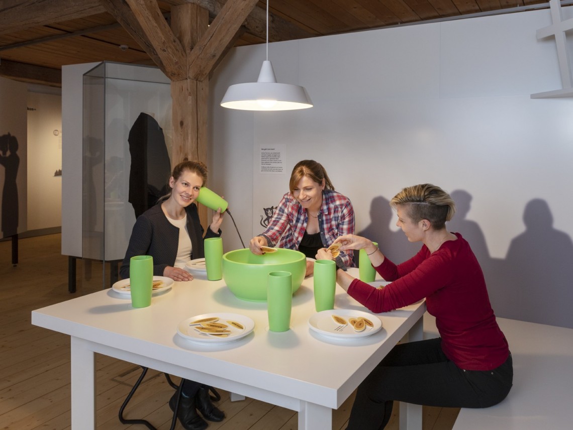 Drei Frauen sitzen um einen weißen Tisch. Darauf stehen Teller, grüne Becher und eine große grüne Schüssel. 