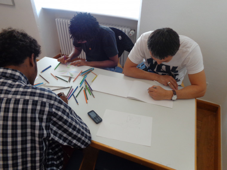 Drei Menschen sitzen an einem Tisch und zeichnen mit farbigen Stiften auf Papier.