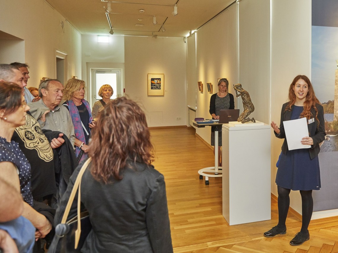 Eine Frau mit einem Blatt in der Hände spricht vor einer Gruppe von Menschen in einem Ausstellungraum. 