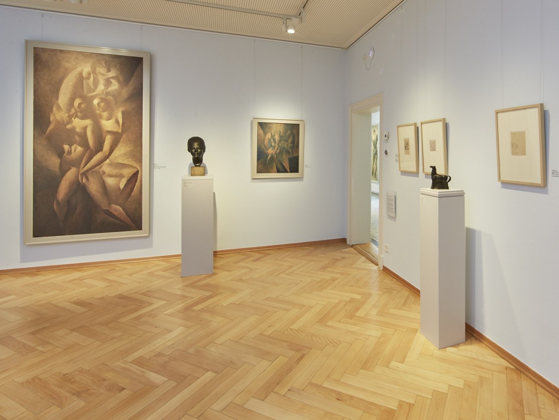 In einem Ausstellungsraum mit Parkettboden sind Bilder an der Wand und zwei Skulpturen auf Sockeln