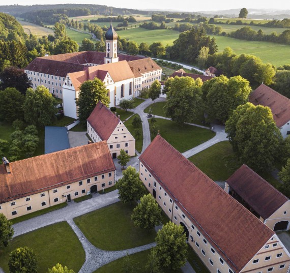 Ein Klostergebäude mit Kirche und Zwiebelturm steht in einer Landschaft mit Wiesen und Hecken.