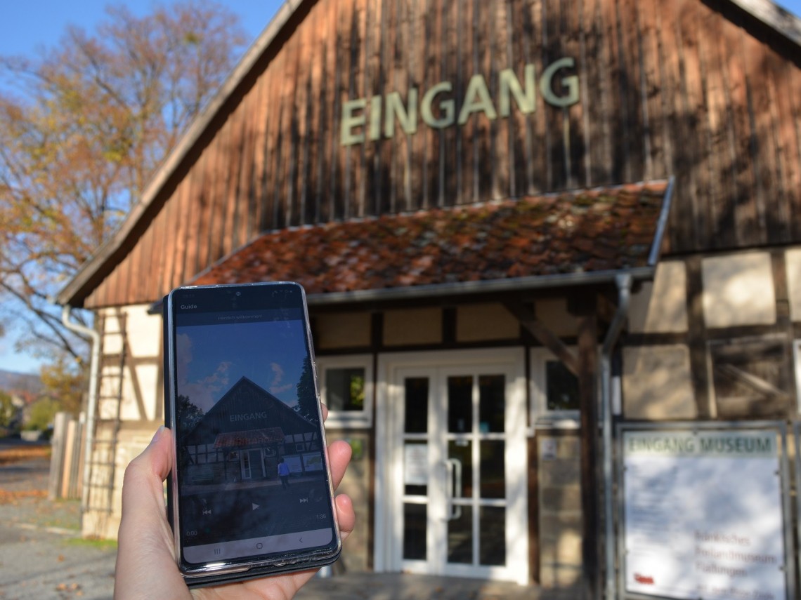 Eine Hand hält ein Smartphone. Im Hintergrund ist ein Fachwerkgebäude über dem die Buchstaben EINGANG angebracht sind.