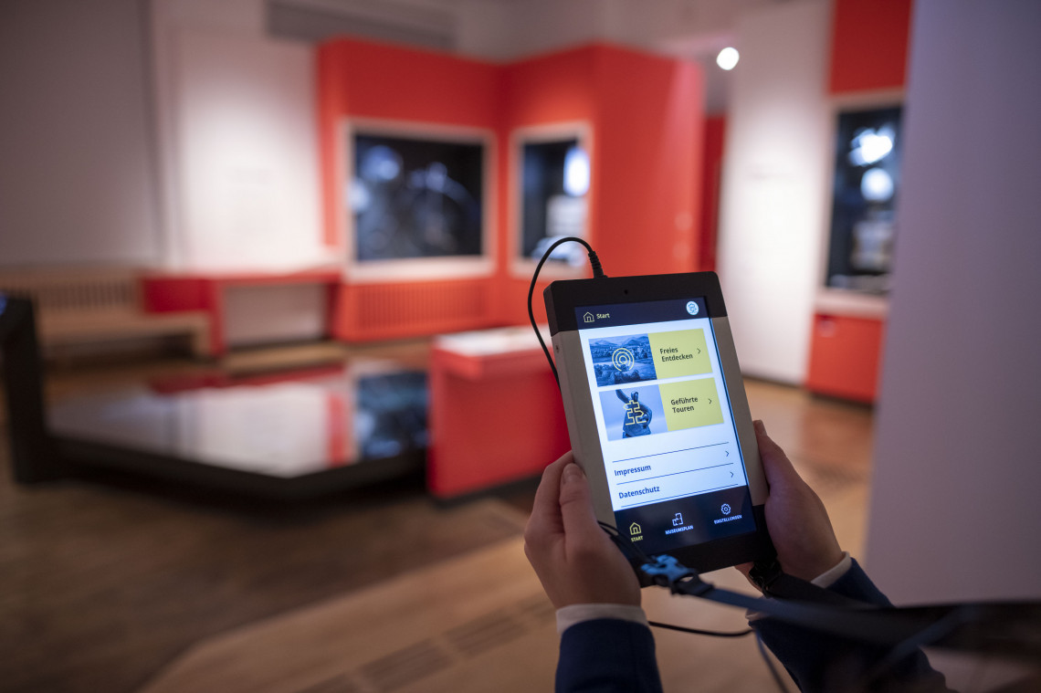 Ein Tablet mit Informationen zur Ausstellung wird von zwei Händen gehalten. Im Hintergrund ist die Ausstellung unscharf zu sehen.
