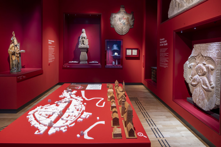 In einem Ausstellungsraum mit roten W&auml;nden stehen verschiedene Figuren an den W&auml;nden. In der Mitte steht ein Stadtmodell auf einem Tisch.