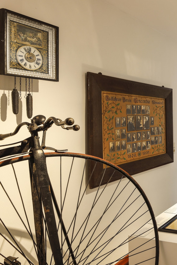 Eine historische Pendeluhr und eine historische Tafel mit Mitgliedern des Radfahrvereins hängen an einer Wand. Davor steht ein Hochrad.