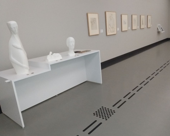 Das Bild zeigt einen weißen Tasttisch mit drei Tastmodellen von einer Skulptur und zwei Plastiken der Würzburger Bildhauerin Emy Roeder. Es sind ganz links „Die Schwangere“ von 1920, in der Mitte die „Ruhende Schaffamilie“ von 1937 und ganz rechts ein Selbstbildnis aus dem Jahr 1964. Vor jedem Tastmodell befinden sich ein QR-Code und ein NFC. Mit einer App kann man hier weitere Informationen zum Leben Emy Roeders und zur jeweiligen Skulptur bzw. Plastik abrufen. Rechts neben dem Selbstbildnis liegen vier Quadrate aus den Materialien, die Emy Roeder für ihre Kunstwerke verwendet hat. Es sind oben links Bronze, oben rechts Kalkstein, unten links Holz und unten rechts Kunststein. Links an der Taststation befindet sich eine Halterung für einen Langstock oder einen Gehstock. Vor dem Tasttisch führt ein taktiles schwarzes Bodenleitsystem an sechs Zeichnungen vorbei zu einer Tastzeichnung ganz hinten im Raum. Vor den Tastskulpturen und der Tastzeichnung befindet sich jeweils ein Aufmerksamkeitsfeld. 
