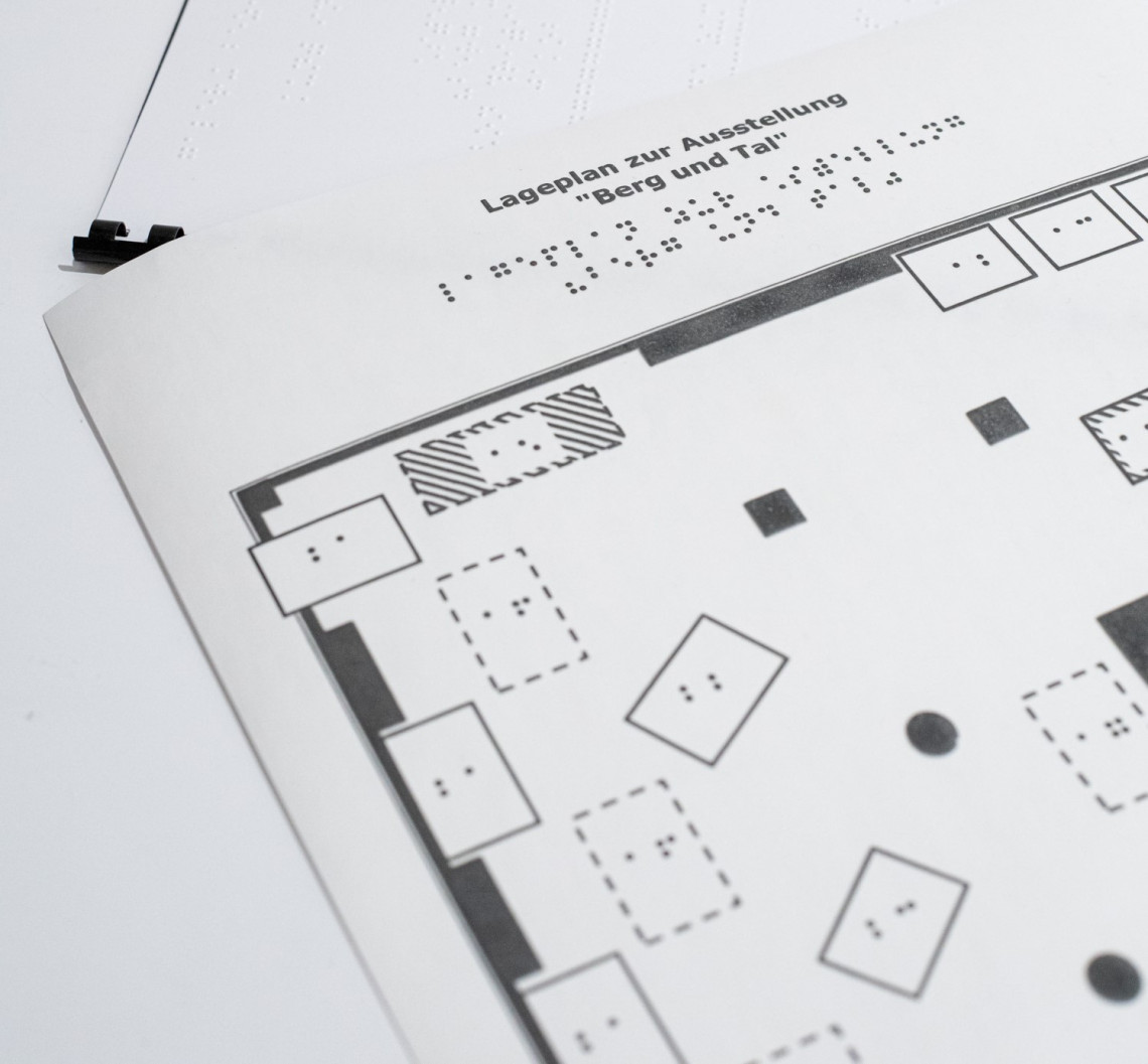 Ausschnitt eines barrierefreien Lageplans zum Tasten mit Nummerierungen in Brailleschrift, sowie im Hintergrund der Ausschnitt eines Brailletextes.